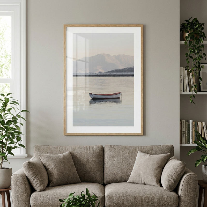 A framed art print depicting a serene lake scene with a small boat and distant mountains, displayed on a light gray wall above a beige sofa in a styled living room.