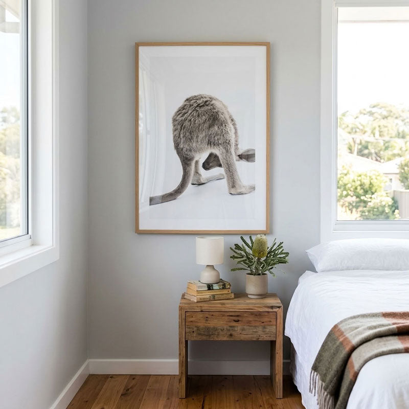 A framed black and white kangaroo art print hangs on a light gray wall in a bedroom setting, above a rustic wooden nightstand with a lamp and plant next to a bed.