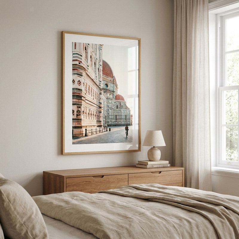 A framed art print depicting a scenic view of Florence's Duomo cathedral is displayed on a bedroom wall above a wooden nightstand with a lamp and books, next to a bed with beige linens.