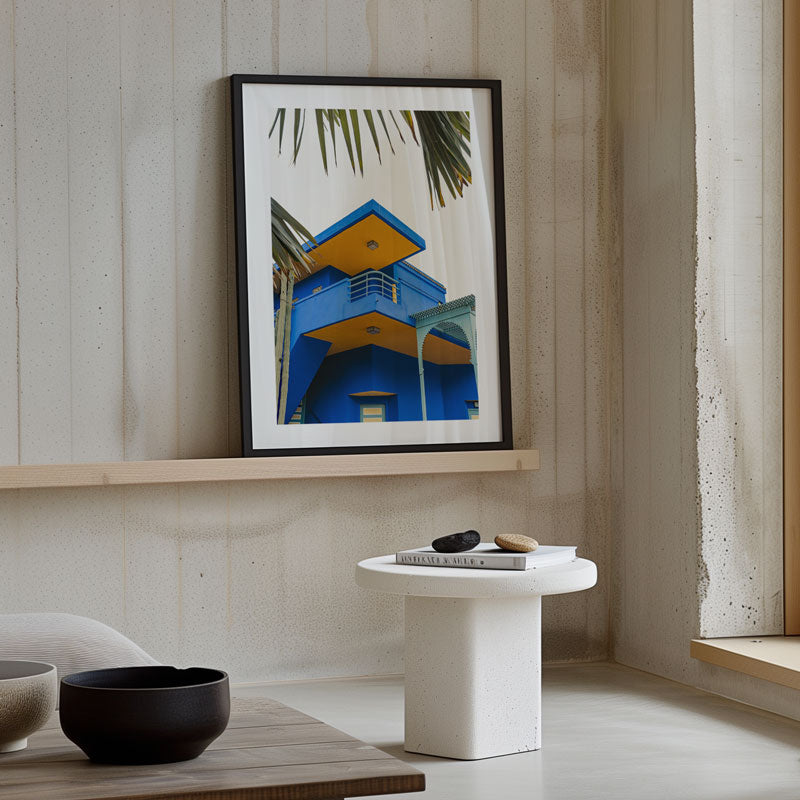 Framed artwork of a blue building with palm trees on a wall above a white table.