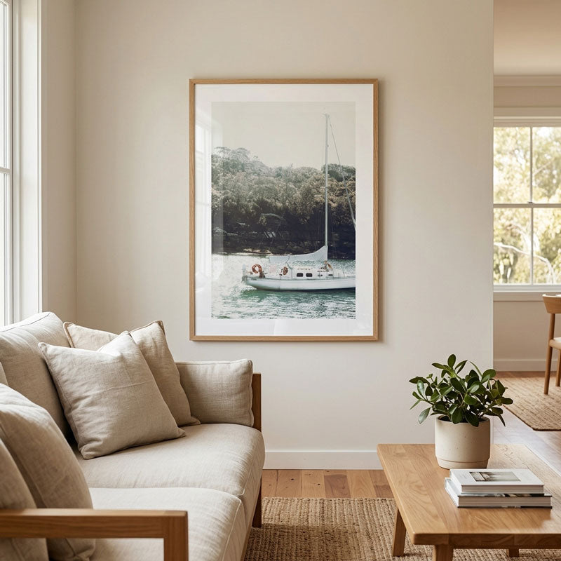 A framed art print depicting a white sailboat on a calm lake surrounded by green trees, displayed on a beige wall in a modern living room setting with a sofa, coffee table, and potted plant.