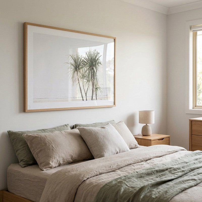 A large framed botanical art print of palm trees hangs on a light beige wall above a neatly made bed with neutral beige and sage green bedding in a softly lit bedroom.