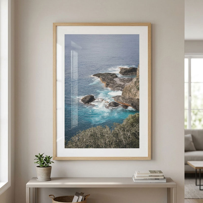 A framed coastal landscape print hangs on a beige wall above a console table. The artwork shows blue ocean water, rocky shoreline, and bright turquoise surf, with a light wood frame and white mat.