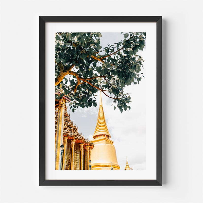 A framed art print featuring a low-angle photograph of a golden temple stupa under a tree with green leaves, presented in a black frame against a white wall.