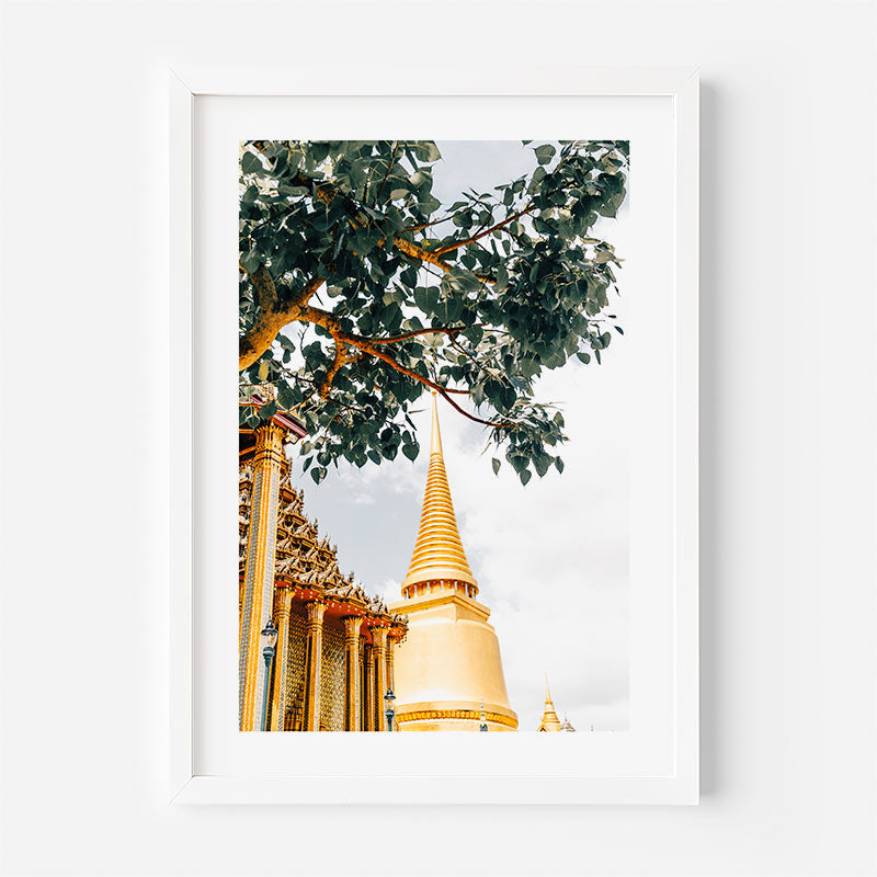 A framed art print featuring a golden temple stupa and ornate columns under a tree with green leaves, displayed on a clean white wall.