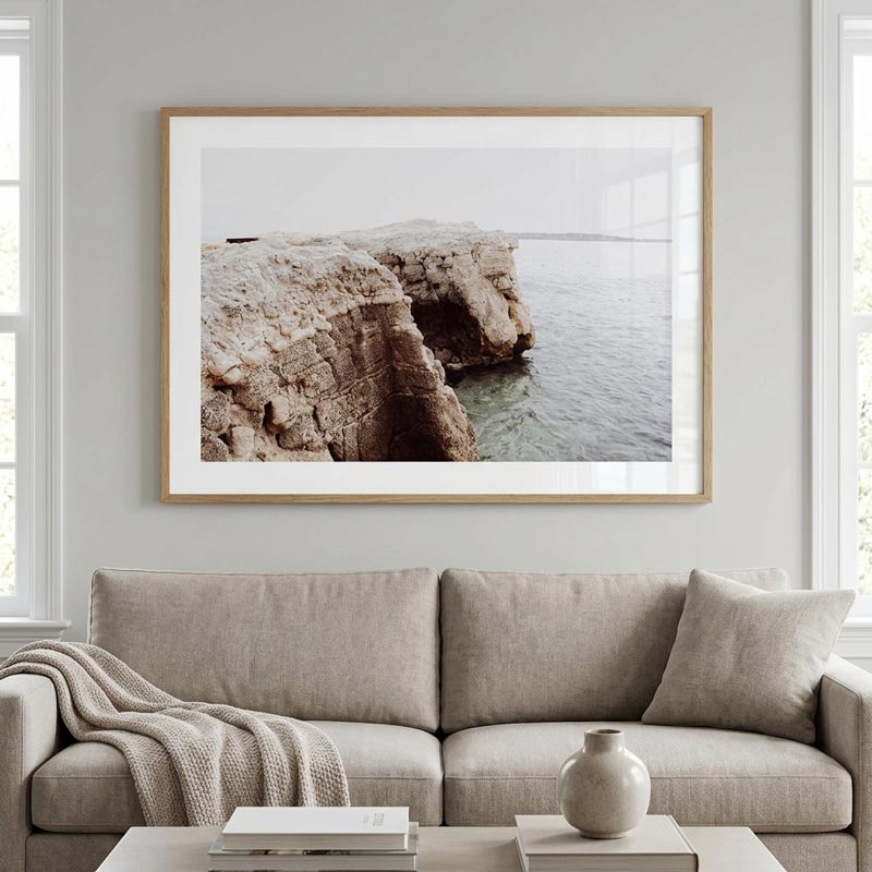A framed landscape art print of coastal cliffs and water hangs on a light gray wall above a beige fabric sofa in a bright, modern living room.