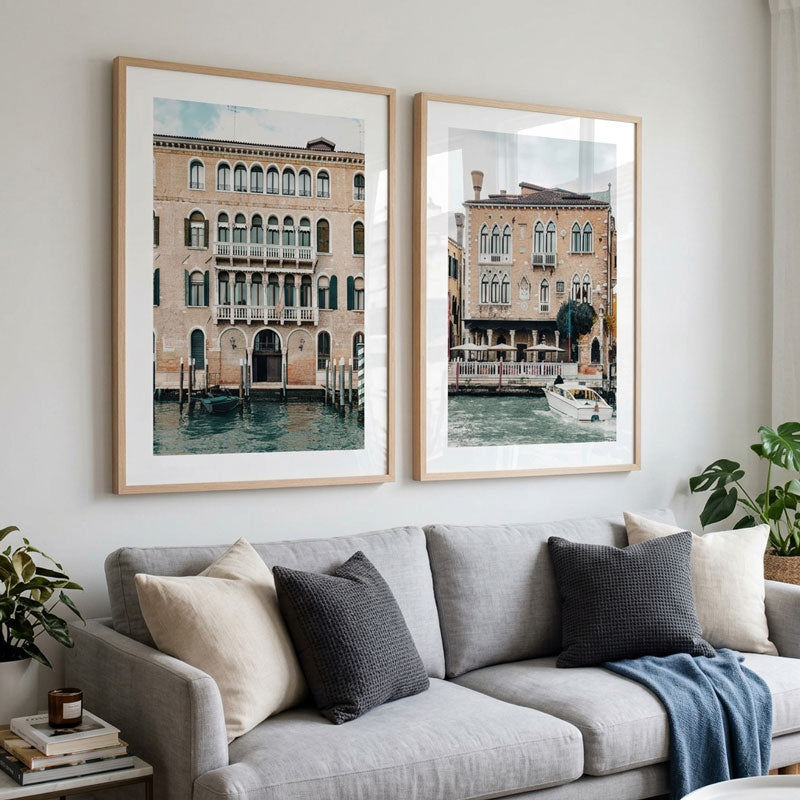 Two framed art prints depicting Venetian canal houses hang on a light gray wall above a modern gray sofa with neutral throw pillows in a styled living room.