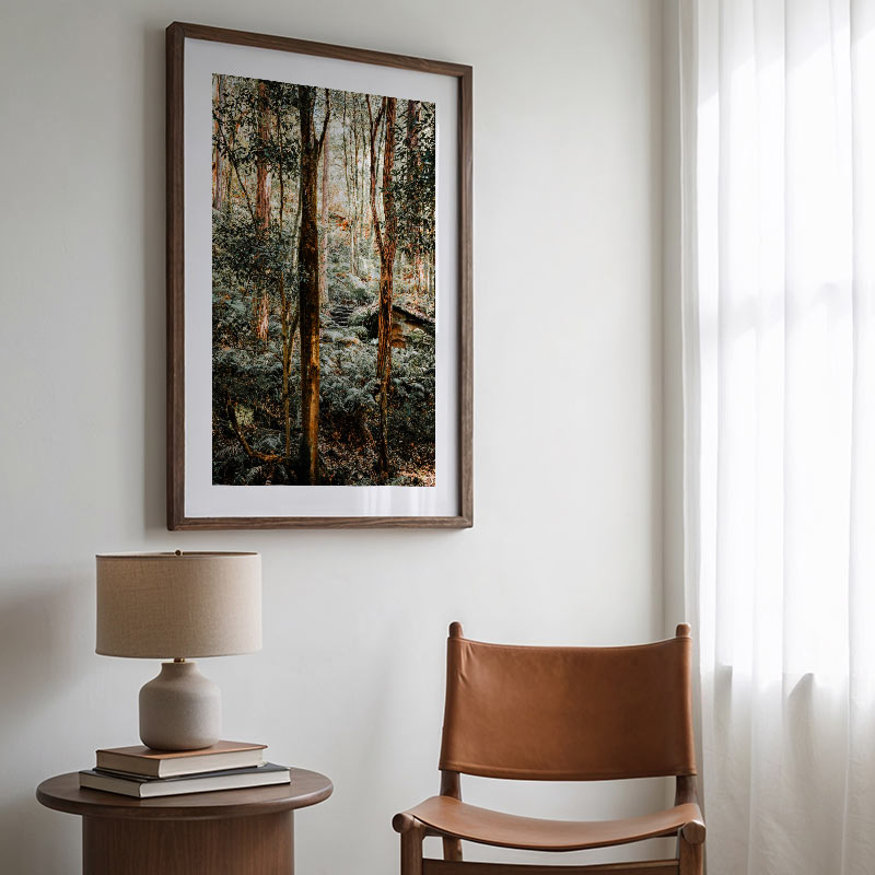 Framed forest artwork on a wall above a table with a lamp and books, next to a brown leather chair.