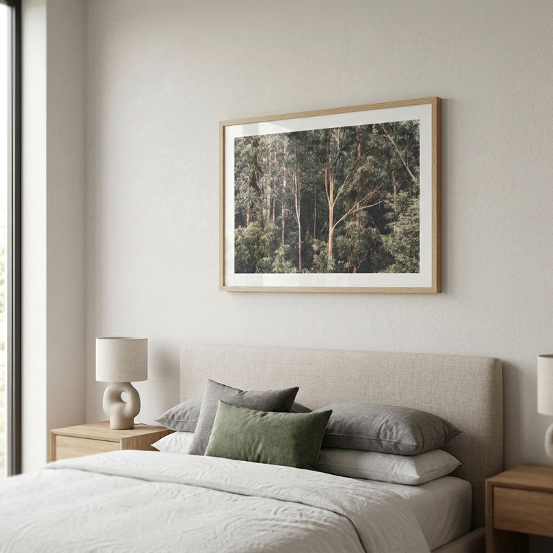 A framed forest art print hangs on a light beige wall above a neatly made bed with neutral-toned bedding and pillows in a modern, minimalist bedroom setting.