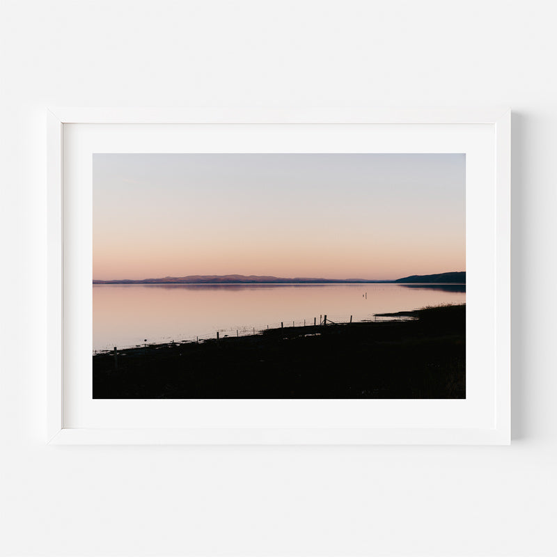 Framed photograph of a sunset over a calm body of water with a silhouette of a fence and landscape.