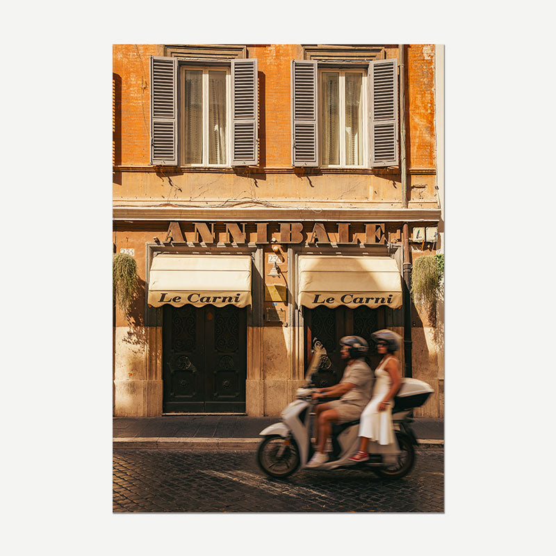 A vertical photographic print of a warm-toned European street scene featuring a building facade with shuttered windows and two awnings reading 'Le Carni', plus a scooter with two riders moving across the cobblestone street. The print is centered on a light gray background.