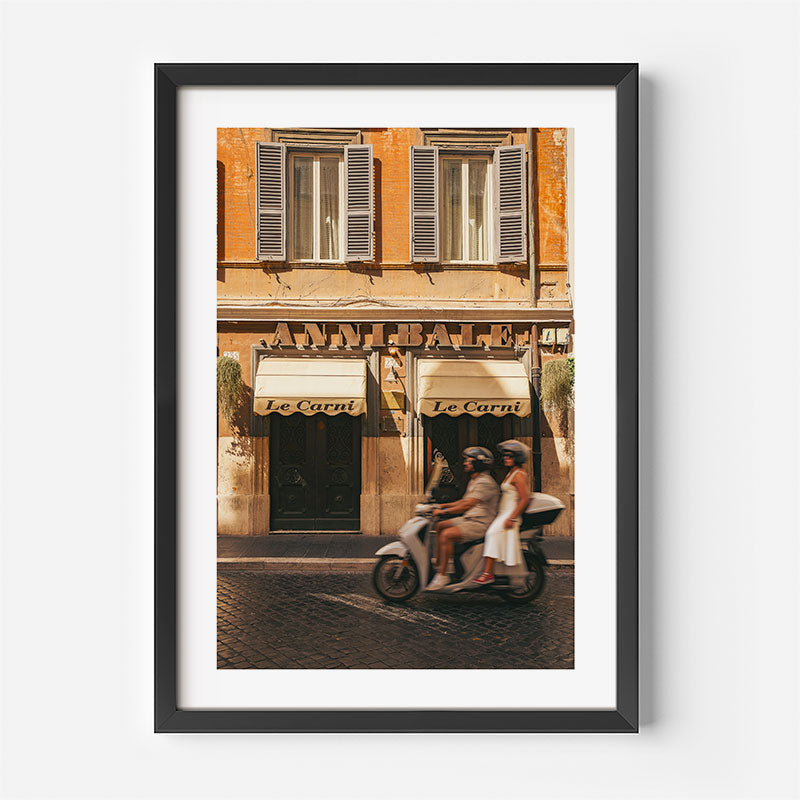 A framed art print featuring a vintage photograph of a warm-toned European building with shuttered windows, awnings reading 'Le Carrot', and a scooter with two riders on a cobblestone street. The print is displayed in a black frame with a white mat against a light gray background.