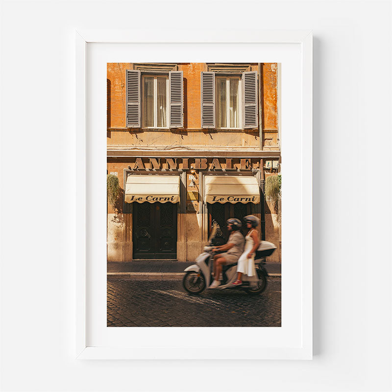 A framed art print featuring a warm-toned photograph of a building facade with shuttered windows and a scooter rider in front of awnings labeled 'Le Carou' and 'Annalet'. The print is presented in a clean white frame against a light gray background.