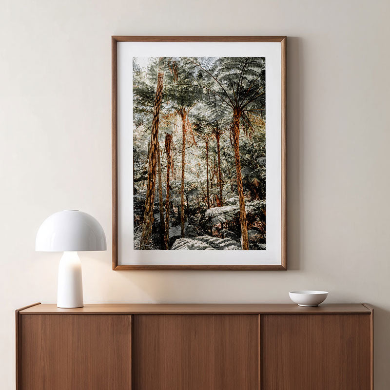 Framed forest print on a wall above a wooden cabinet with a white lamp and bowl.