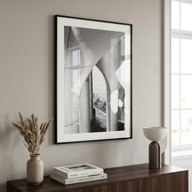 A framed black and white architectural art print with a white mat and black frame, hanging on a light gray wall above a dark wood console table with books, a vase, and a lamp.