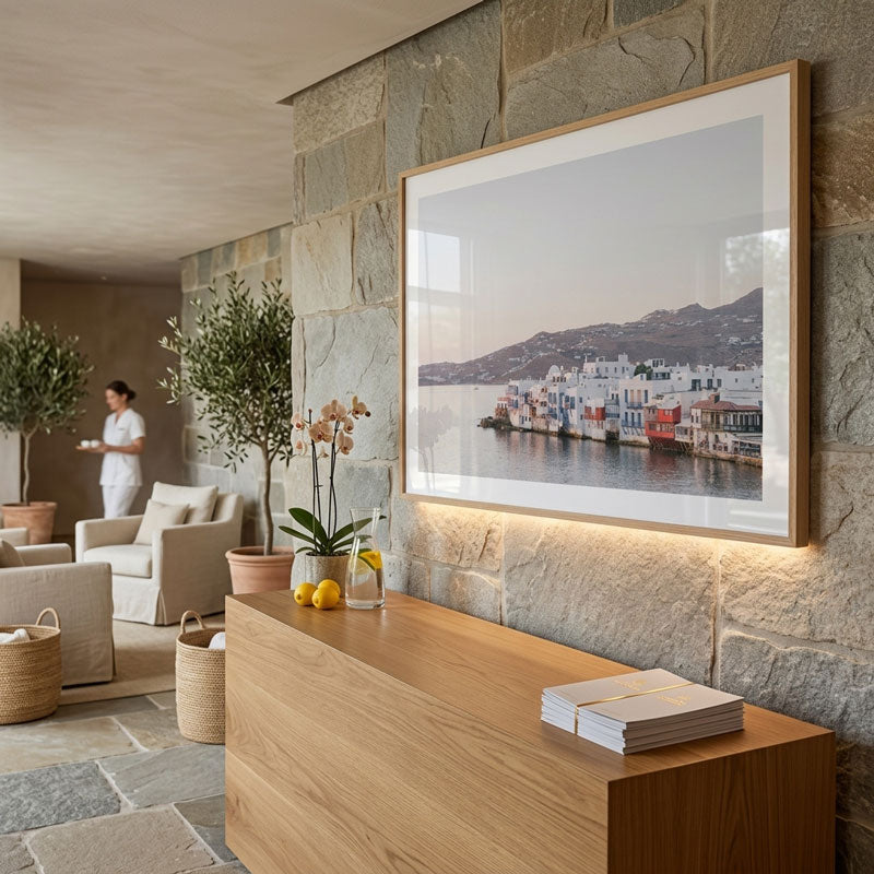 A large framed art print depicting a coastal town with white buildings and colorful boats, displayed on a stone wall above a wooden console table in a modern, sunlit interior.