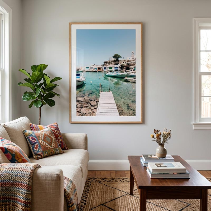 A large framed art print of a coastal harbor with turquoise water, boats, and a wooden pier, displayed on a light gray wall in a styled living room with a sofa, pillows, and a coffee table.