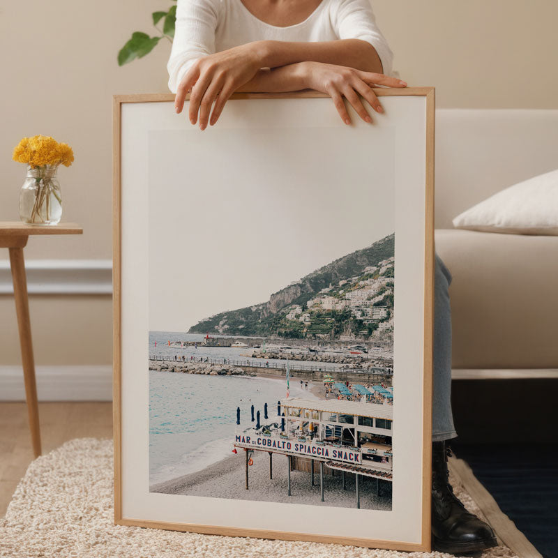 Person holding a framed photograph of a coastal scene with mountains and a pier.
