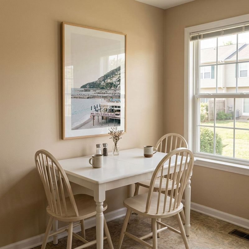 A framed coastal landscape print hangs on a beige wall above a white dining table with four light wood spindle-back chairs in a bright, neutral-toned room.