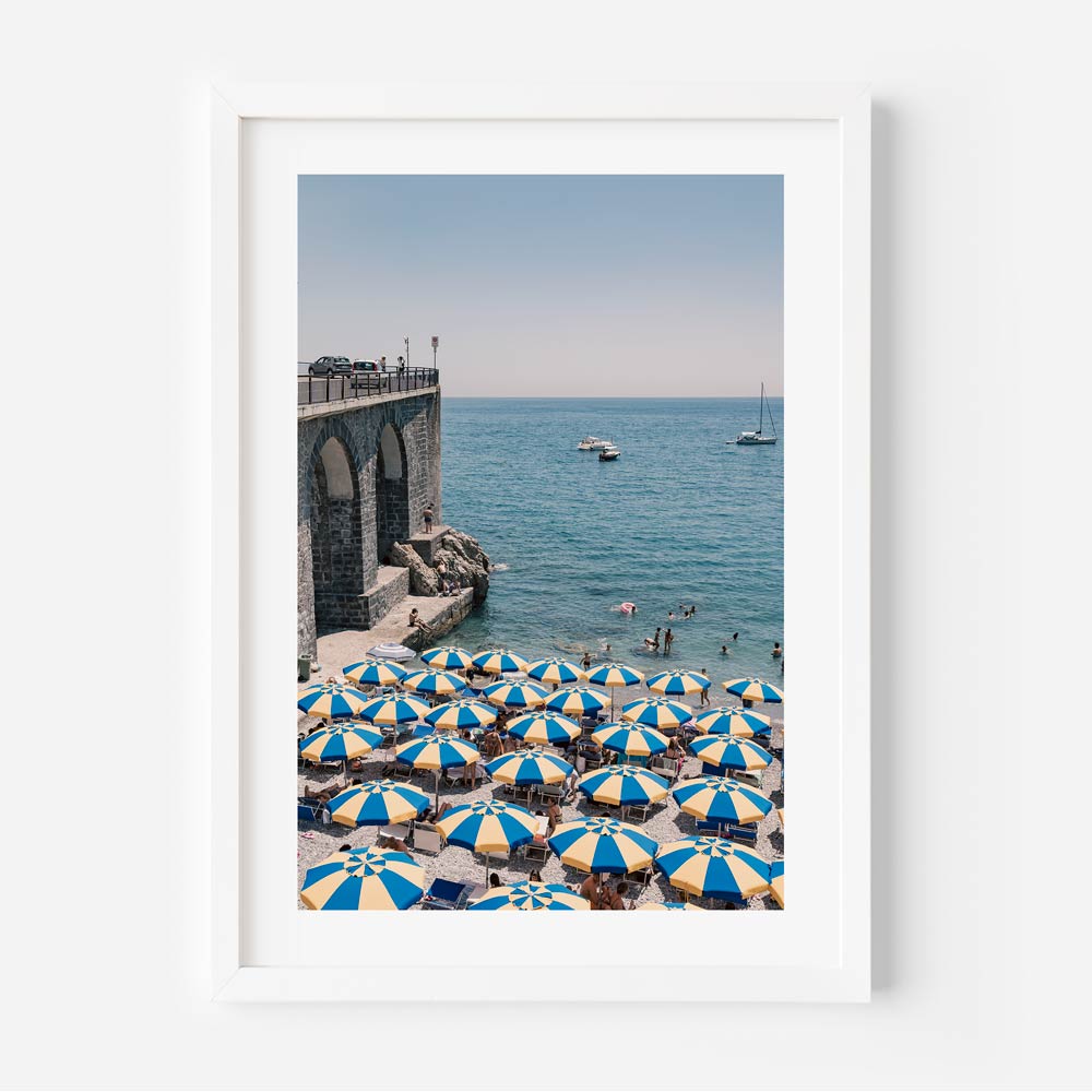 A framed photograph featuring a coastal scene with blue and yellow beach umbrellas and a backdrop of a clear blue sky and calm sea.