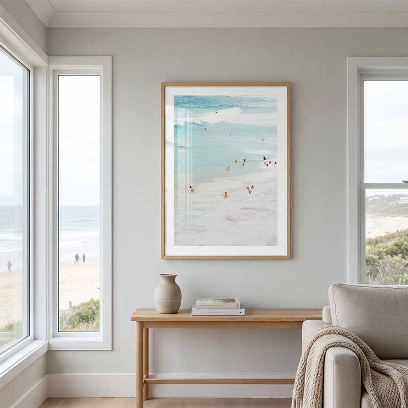 A framed beach art print hangs on a light gray wall above a wooden console table in a bright, modern living room. The print depicts a soft blue ocean and white sand beach with small figures of swimmers.
