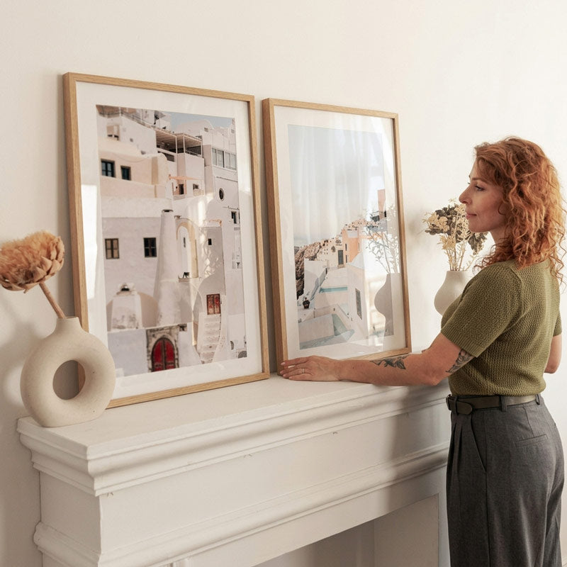 A woman with curly red hair stands beside a white mantel, holding a second framed art print next to a larger framed print of white buildings with blue accents. The room has a warm neutral color palette with a white mantel and wall, and decorative dried flowers in a vase.