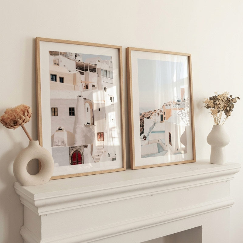 Two framed minimalist architectural art prints with light wood frames, displayed side-by-side on a white mantelpiece decorated with ceramic vases and dried flowers.