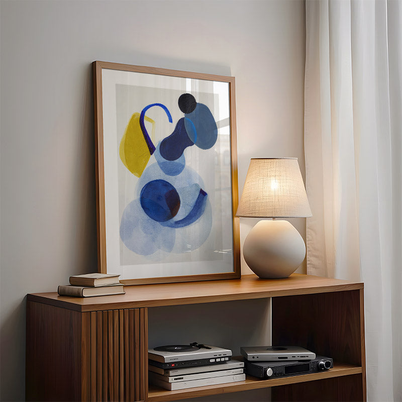 Framed abstract art on a wooden shelf with a lamp and books in a room.