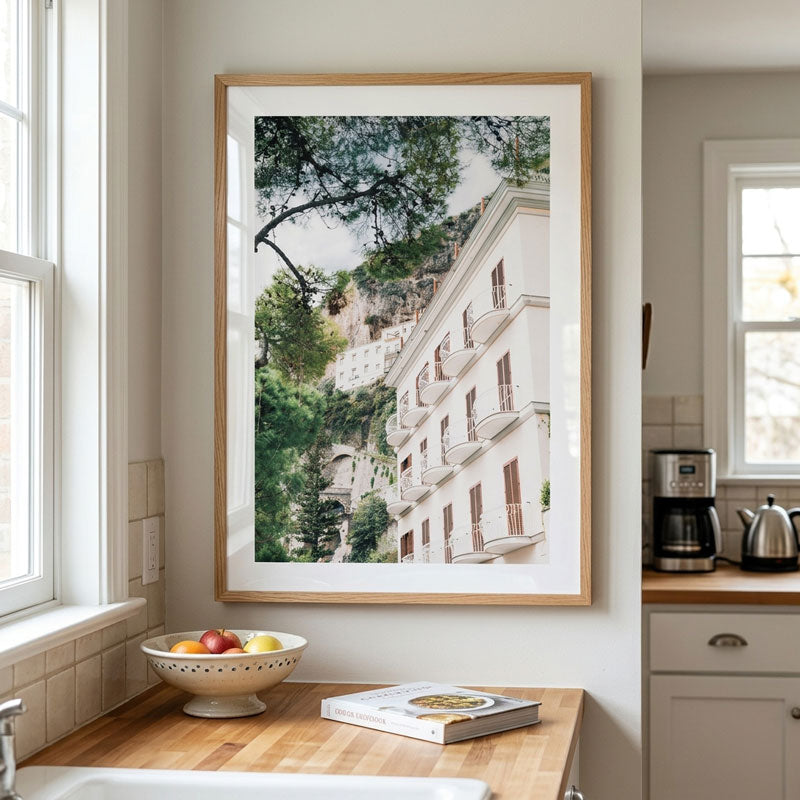 A framed vertical art print depicting a Mediterranean-style white building with balconies and green trees, hanging on a kitchen wall above a wooden countertop.