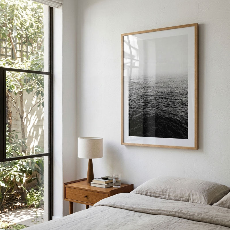 A framed black and white photograph of ocean waves hanging on a white bedroom wall above a wooden nightstand and bed.