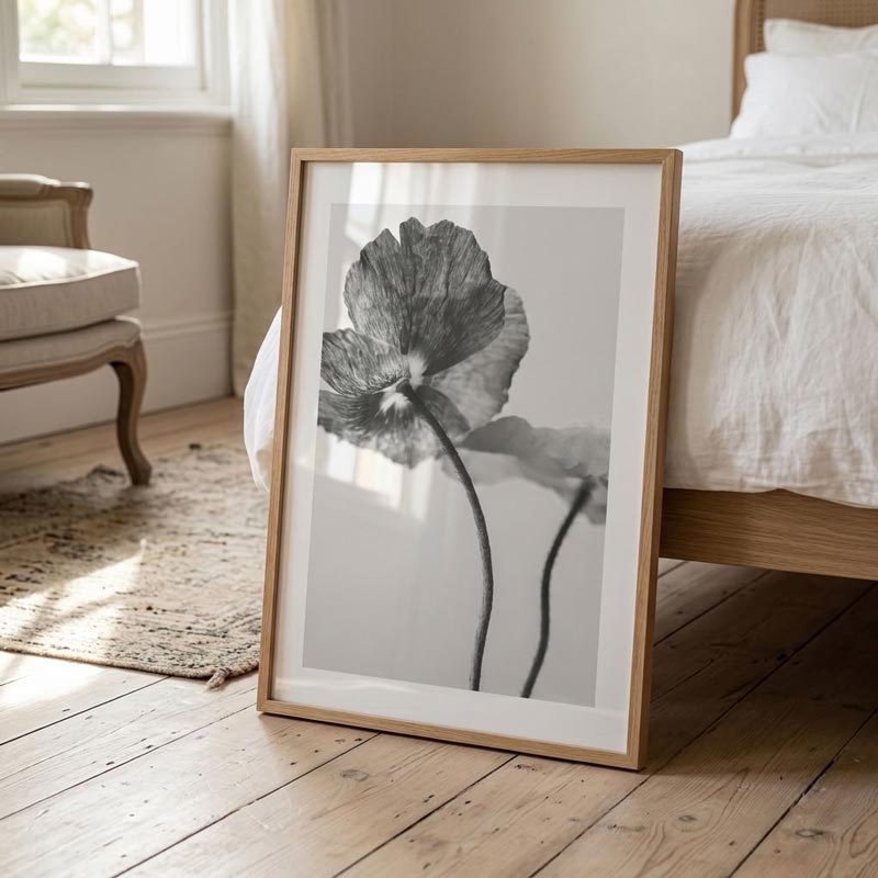 A framed black and white botanical art print featuring a poppy flower, leaning against a bed in a softly lit bedroom with light wood floors and white bedding.