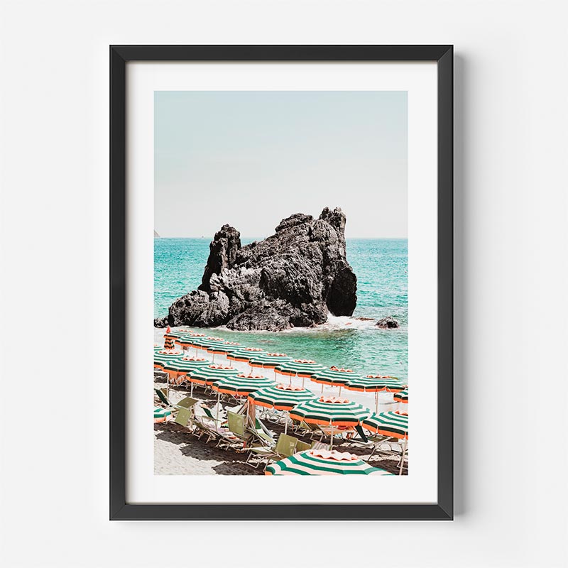 Framed artwork of a beach scene with lounge chairs and umbrellas near a rock formation.