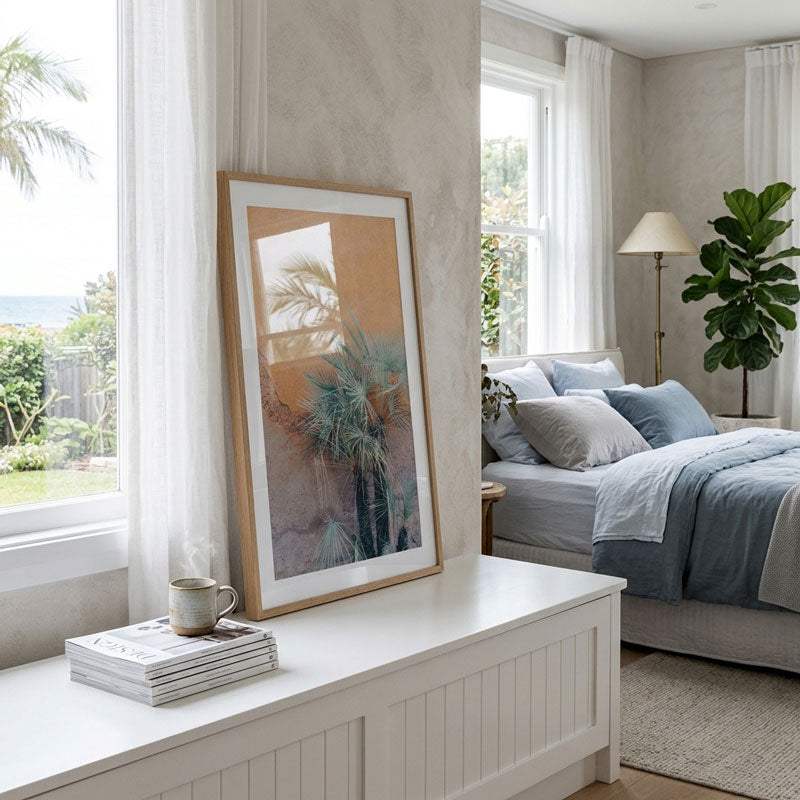 A large framed art print featuring a warm-toned palm tree illustration in a light wood frame, styled on a white bench in a bright, modern bedroom.