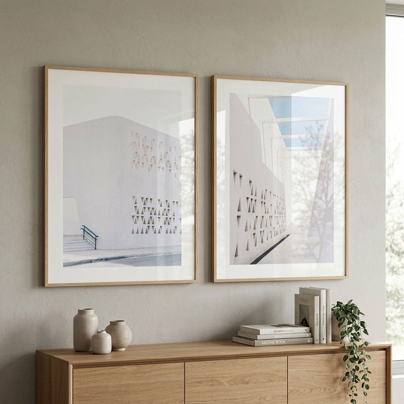 Two framed minimalist architectural art prints hang on a light gray wall above a wooden console table with decorative vases, books, and a trailing plant.