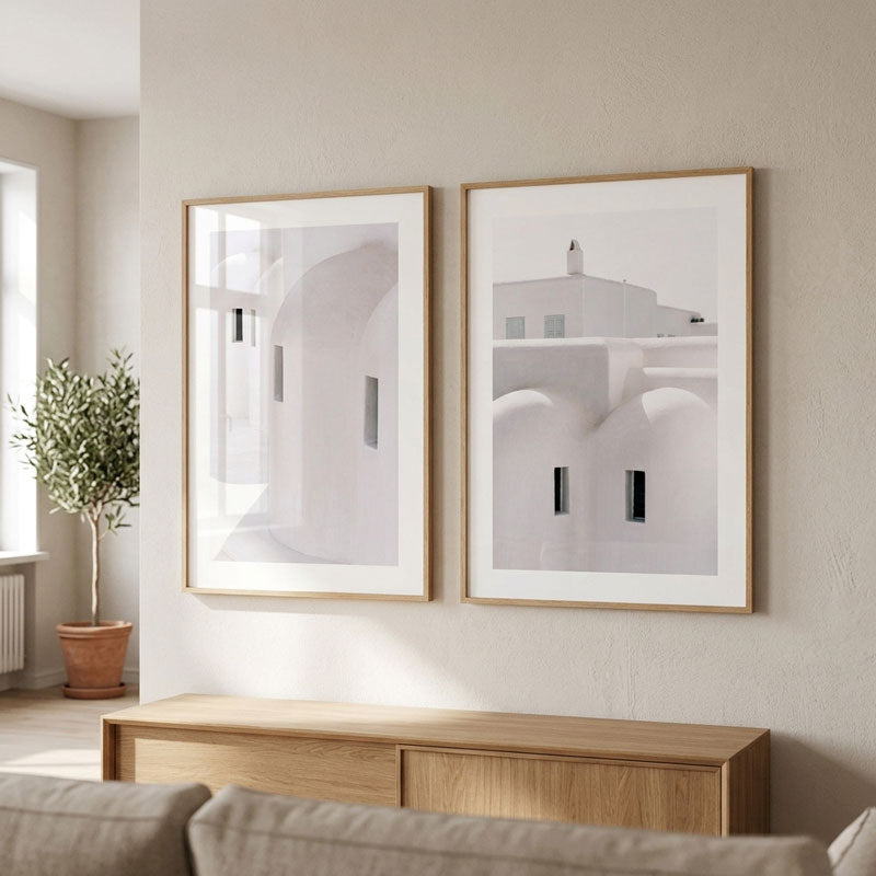 Two framed minimalist architectural art prints with white and gray tones and light wood frames, displayed side-by-side on a beige wall above a wooden console table in a modern living room setting.