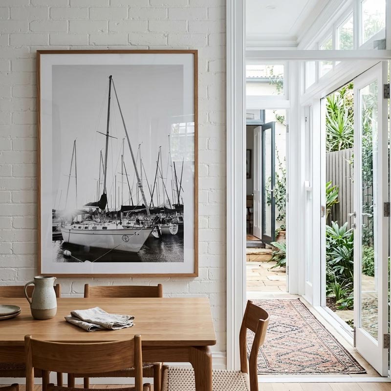 A framed black and white photograph of sailboats hanging on a white brick wall above a wooden dining table in a bright, modern room with an open glass door leading to a garden patio.
