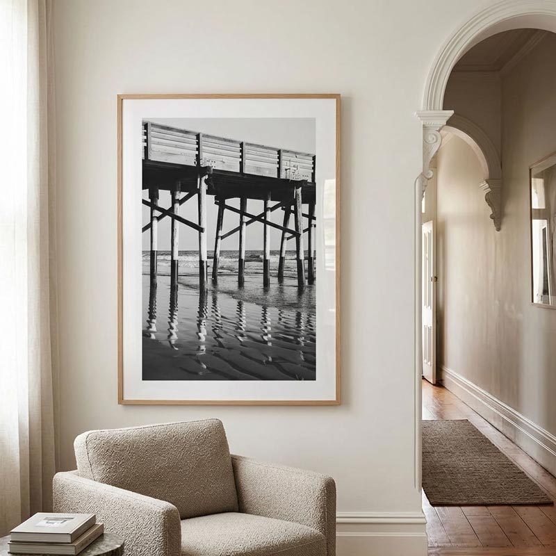 A framed black and white photograph of a pier over water, displayed on a cream wall in a modern living room setting with a textured armchair and an arched hallway.