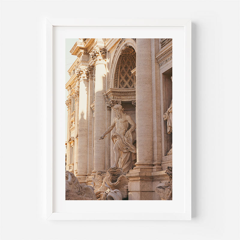 A framed art print featuring a photograph of the Trevi Fountain in Rome, showcasing the white marble statue of Neptune and the warm beige stone architecture. The print is presented in a clean white frame against a light gray wall.