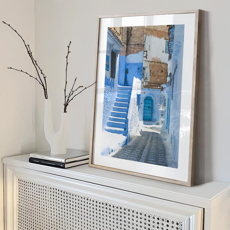 Framed artwork of a blue street scene on a white console table with a vase and books.