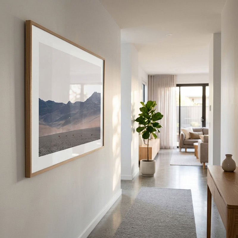 A large framed landscape art print featuring a mountain range, displayed on a white wall in a modern, sunlit hallway with a wooden console table and a potted plant.