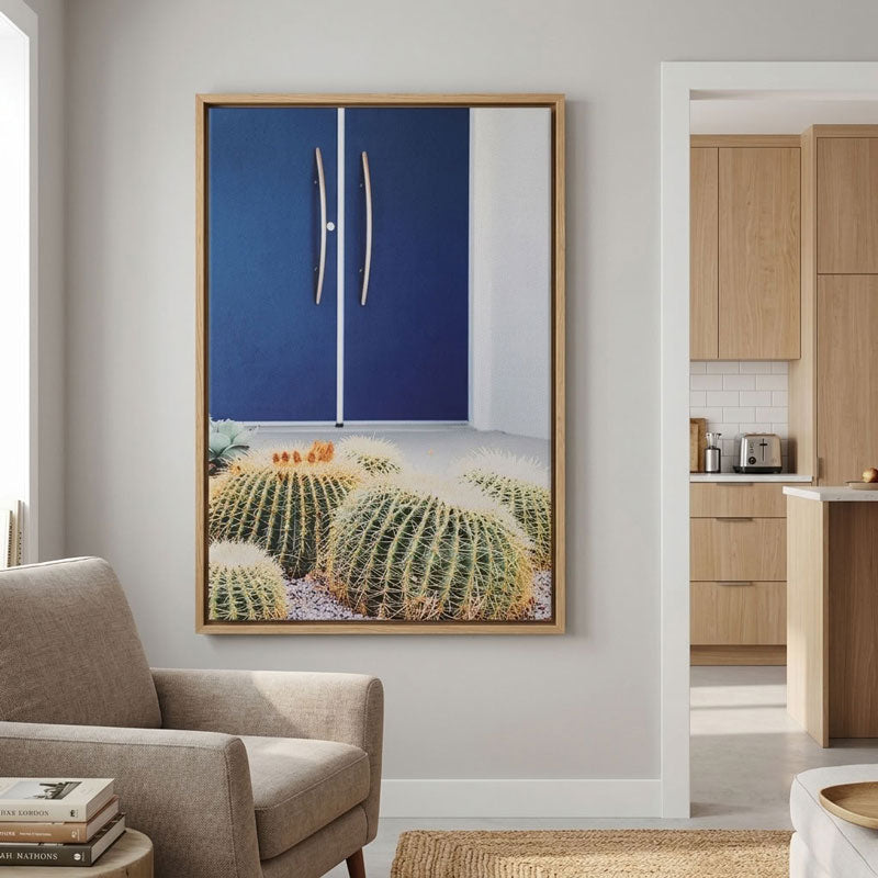 A framed wall art print hanging in a modern living room. The artwork depicts a blue double-door cabinet above a desert scene with barrel cacti.