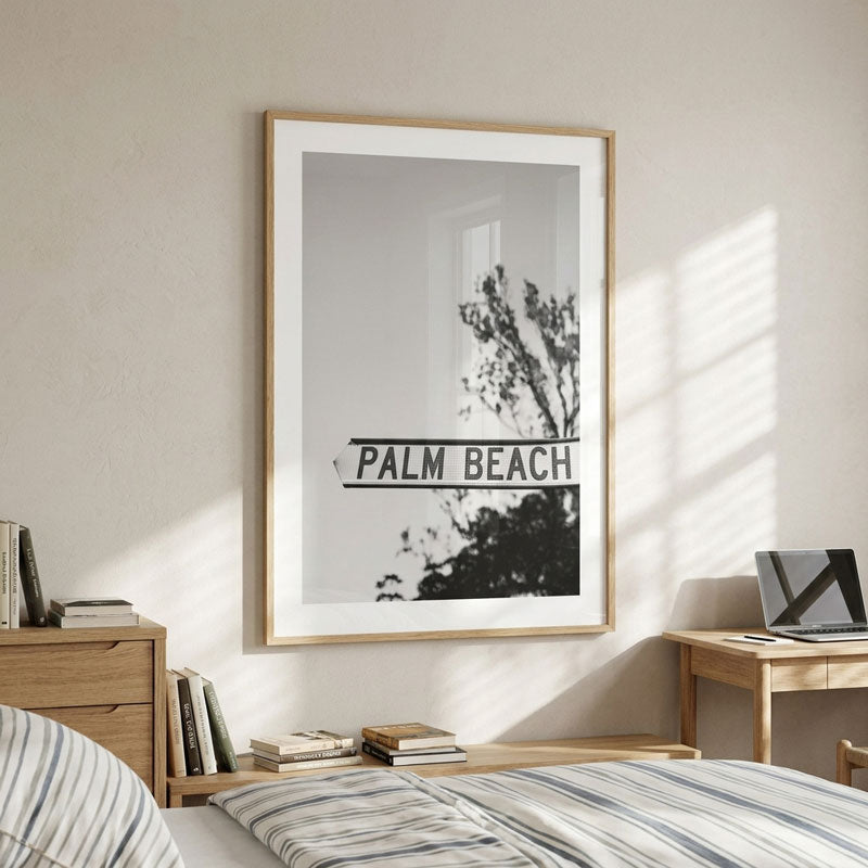 A framed black and white art print featuring a street sign that reads 'PALM BEACH' with tree branches, displayed on a beige wall above a bed in a modern, sunlit bedroom.