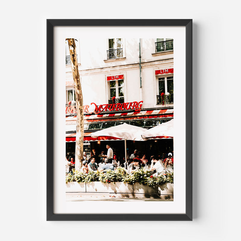 Framed photograph of a bustling outdoor cafe with people sitting under red umbrellas in front of a building with a visible sign.