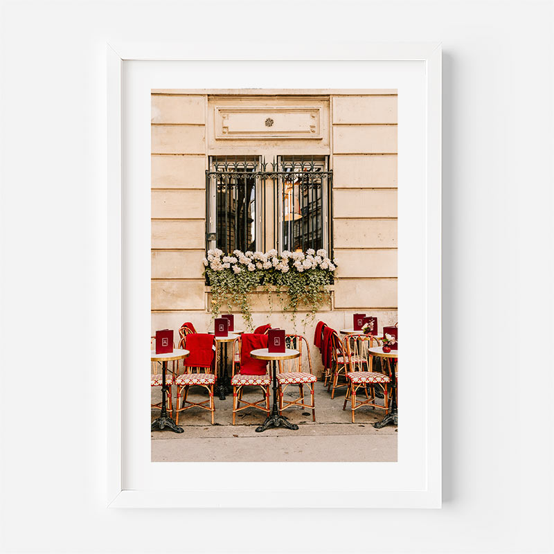 Framed photograph of an outdoor cafe with red chairs and tables against a beige building.