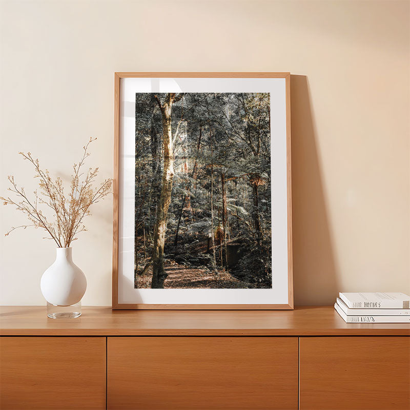 Framed forest print on a wooden shelf with a vase and books.