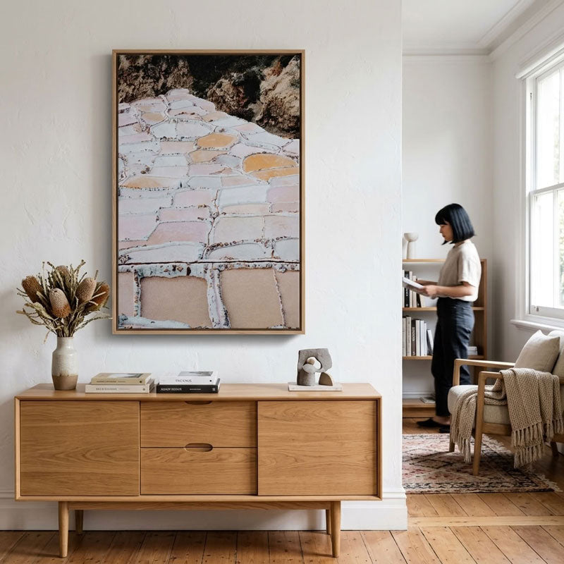 A large framed abstract landscape painting hangs on a white wall above a light wood sideboard in a bright, modern room. A woman is visible in the background near a window and bookshelf.