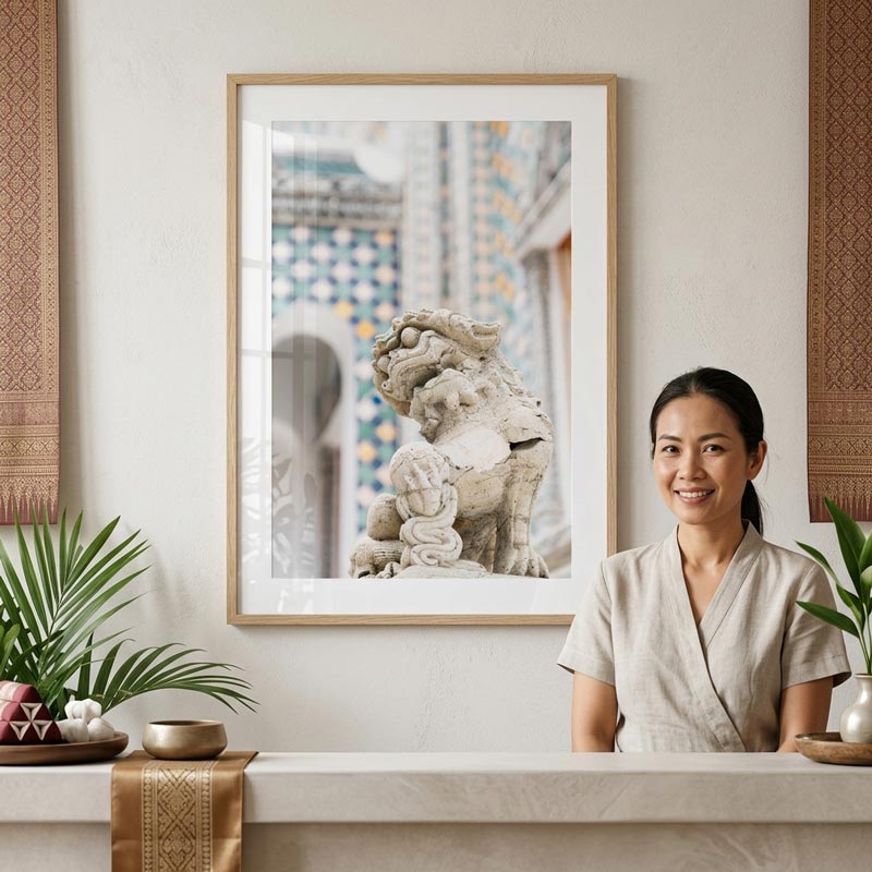 A framed art print featuring a stone lion statue and tiled architecture, displayed on a wall above a counter in a styled indoor setting with a woman seated to the right.