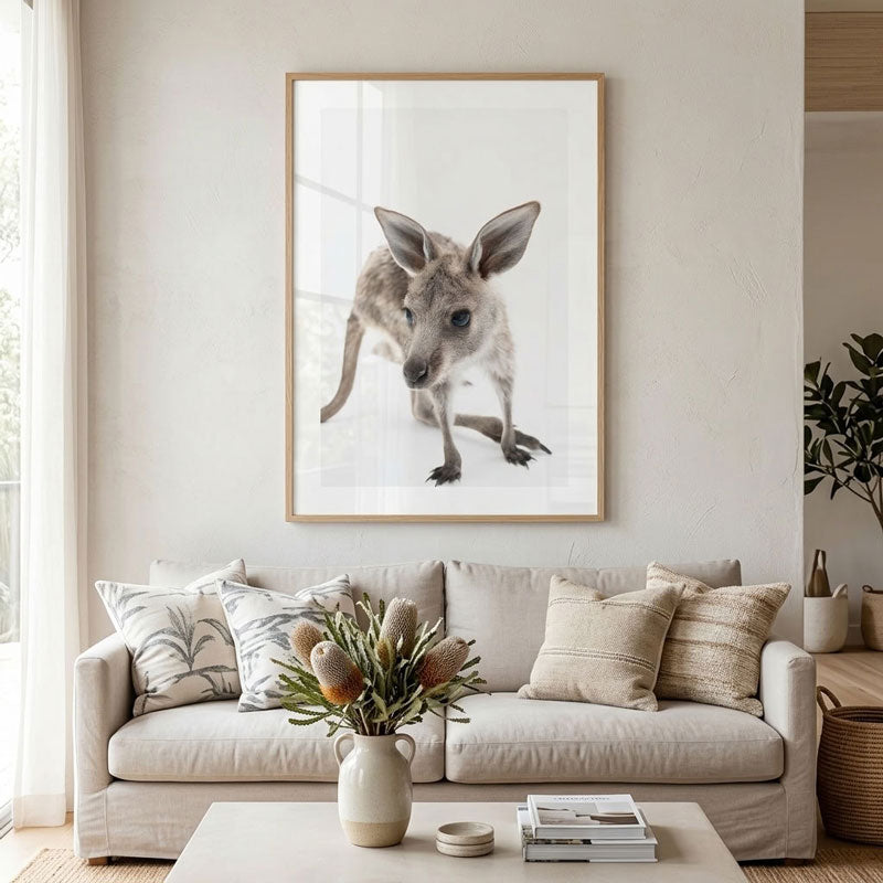 A framed wall art print of a kangaroo hanging above a beige sofa in a bright, modern living room. The print features a realistic close-up photograph of a kangaroo in a light wood frame.