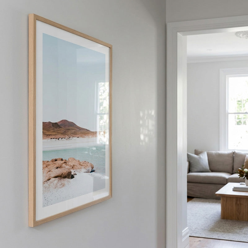 A framed landscape art print hangs on a light gray wall in a modern home interior, showing a beach scene with turquoise water, white sand, and brown rocks. The print has a wide white mat and a light natural wood frame.