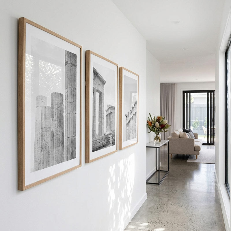 Three framed black and white architectural art prints with light wood frames hang on a white wall in a modern hallway, with a living room visible in the background.